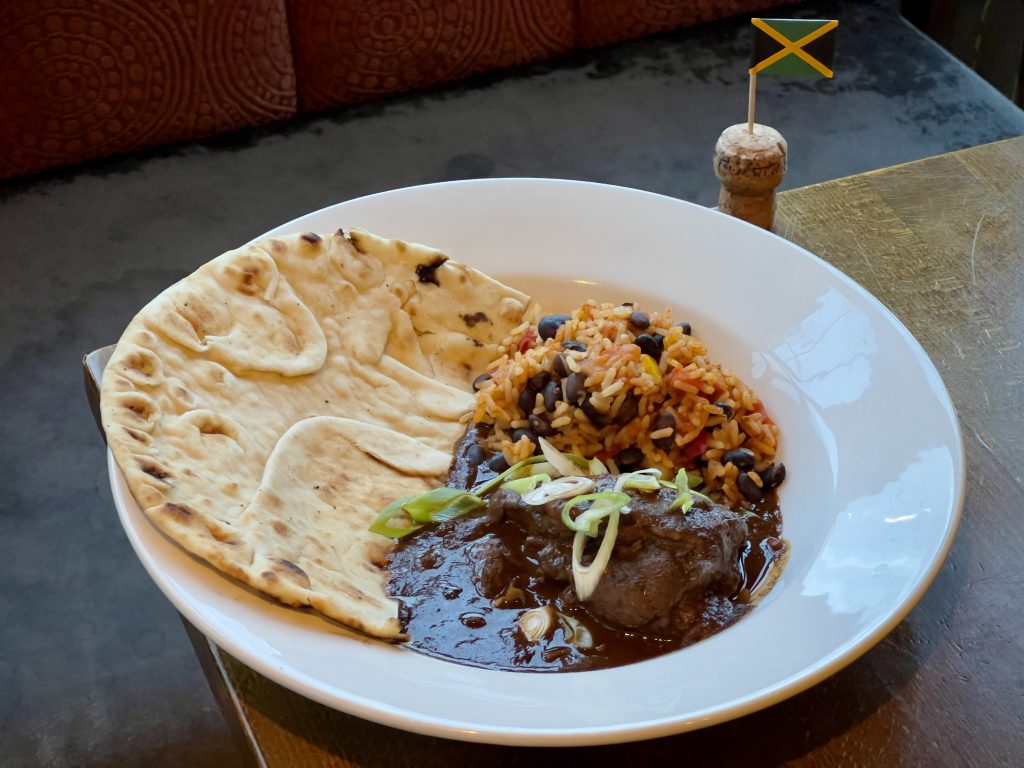A brown stew chicken dish, complete with rice and flatbread, next to a Jamaican cocktail stick flag.