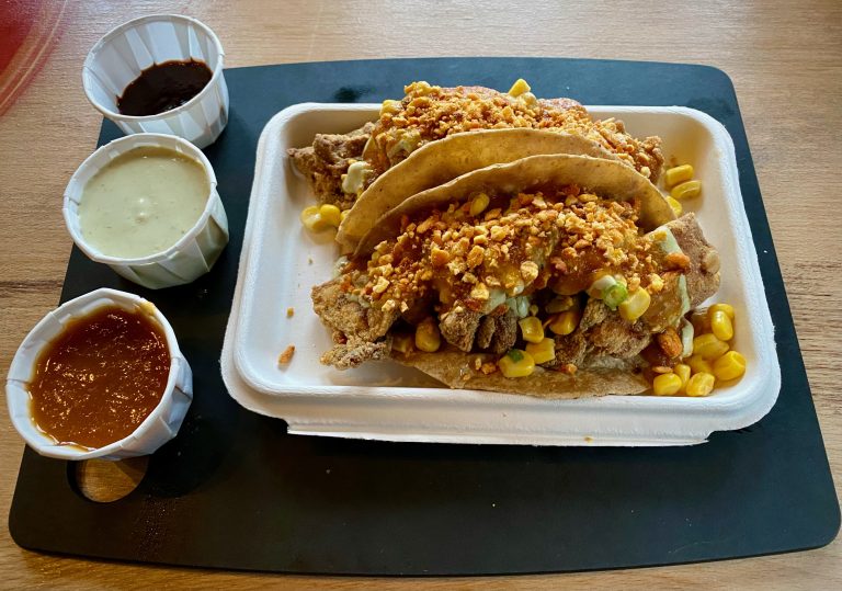 Two fried chicken tacos on a white takeaway tray, served with three different salsas in white takeaway pots.