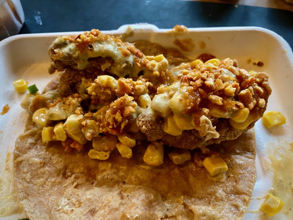 Detail view of a fried chicken taco, comprising fried chicken chunks, sweetcorn pieces and baked corn crumbs, all on top of a corn tortilla.