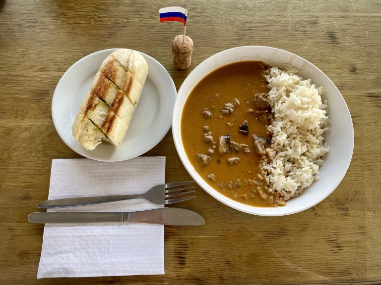 Beef stroganoff and basmati rice in a bowl, a garlic bread baguette on a plate, a wine cork, Russian flag cocktail stick, and cutlery.