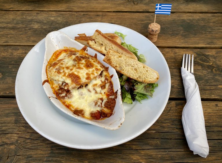 Moussaka, garlic bread and salad on a white plate. The plate is on a wooden table, next to a set of cutlery, and a cocktail stick Greek flag.