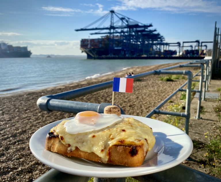 A white plate with a croque madame and French flag cocktail stick on it, resting on a railing. There is a large container ship in a port in the background.
