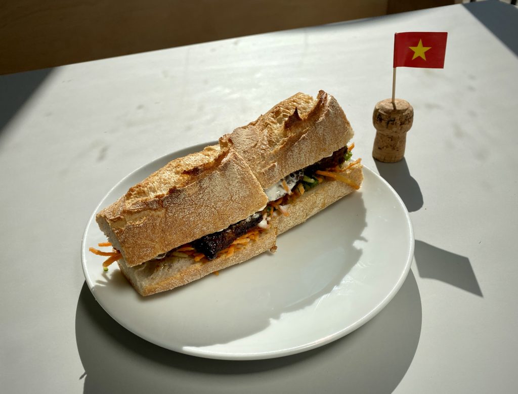 A banh mi containing tofu, carrot and mayonnaise on a white plate next to a Vietnamese cocktail flag.