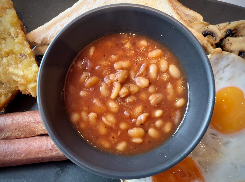A ramekin of baked beans, on a plate with various other Full English Breakfast items.
