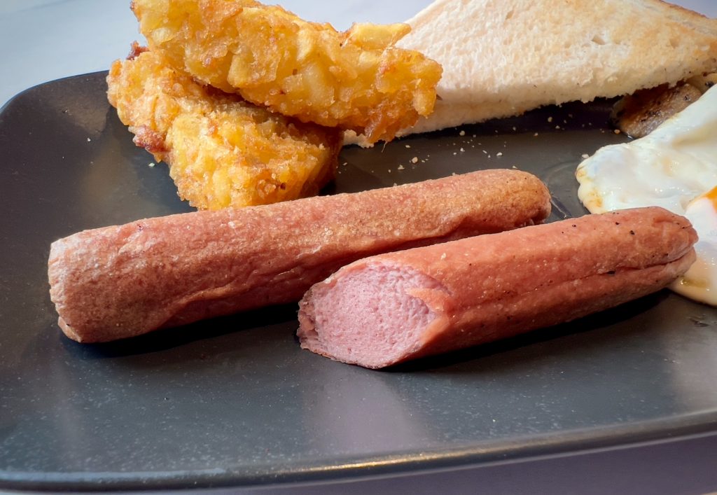 Two sausages on a plate with various other Full English Breakfast items, with one sausage cut in half to show the cross-section.