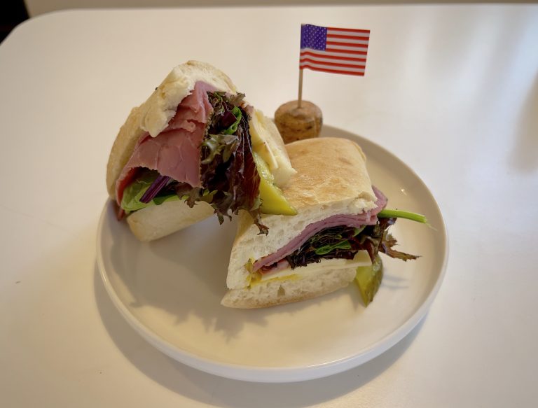 A New York Deli sandwich, complete with pastrami, cheese, mixed leaves and mustard on a plate next to a USA cocktail stick flag.