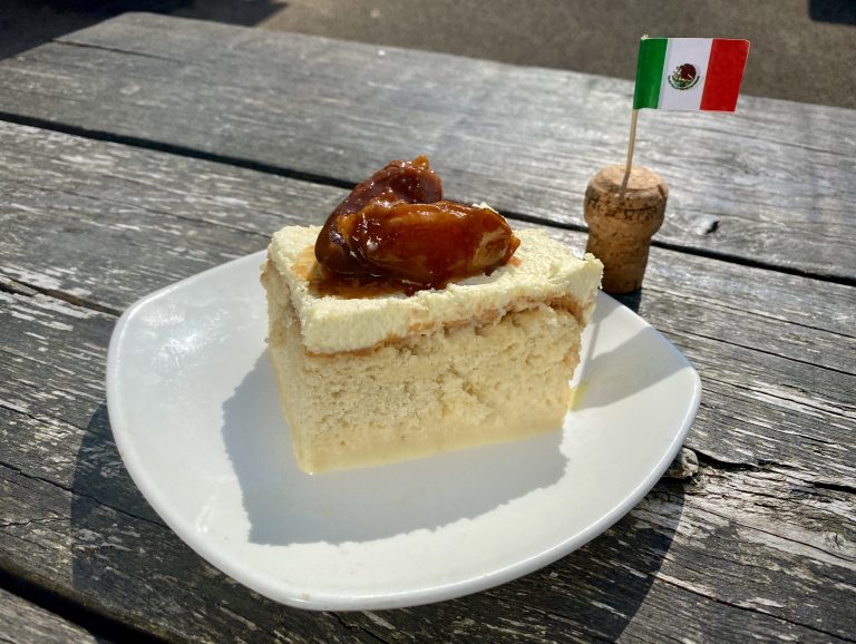 A 'Tres Leches' Mexican pudding on a white plate next to a Mexican cocktail stick flag.