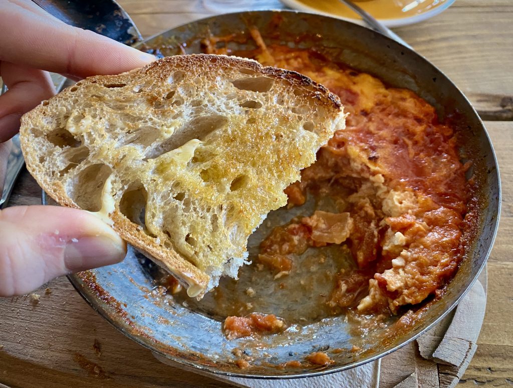 A slice of buttered sourdough toast held in the foreground, with Menemen in a small frying pan in the background.