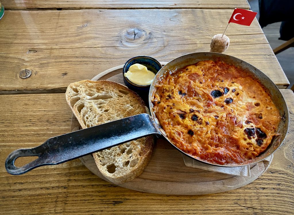 A Menemen in a small frying pan on a wooden chopping board, next to a slice of sourdough toast and butter. There is a Turkish cocktail stick flag on the table.