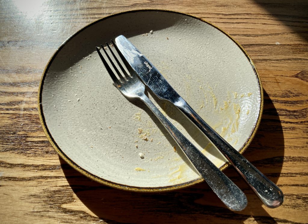 A grey, empty plate on a wooden table, with the knife and fork positioned to show one hand finished eating.
