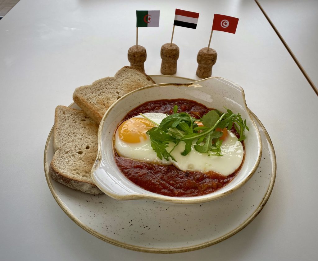 A Shakshuka on a serving platter next to slices of toast and Tunisian, Yemeni and Algerian cocktail stick flags.