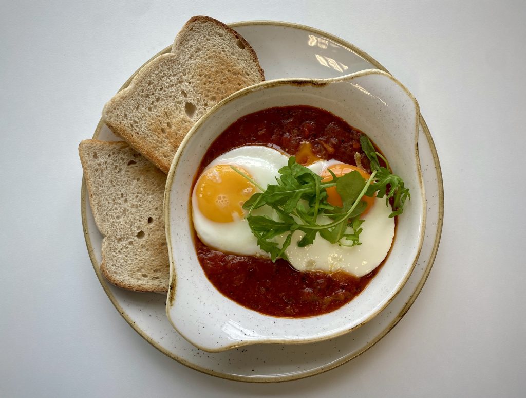 A top-down view of a Shakshuka in a serving platter, next to slices of toast on a plate.