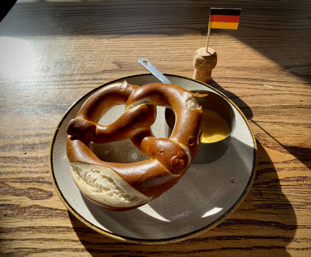 A German Pretzel on a plate with beer cheese sauce, next to a German cocktail stick flag.