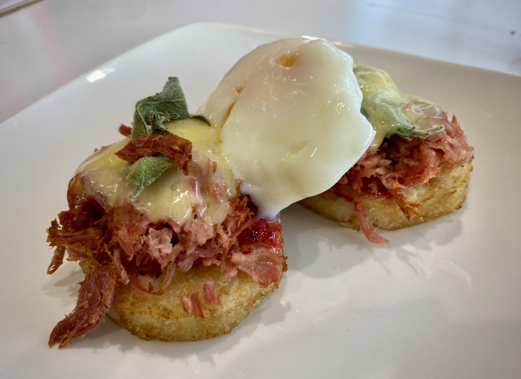 Two potato röstis on a white plate. Ham hock, melted cheese, sage leaves and a poached egg have been placed on top of the röstis.