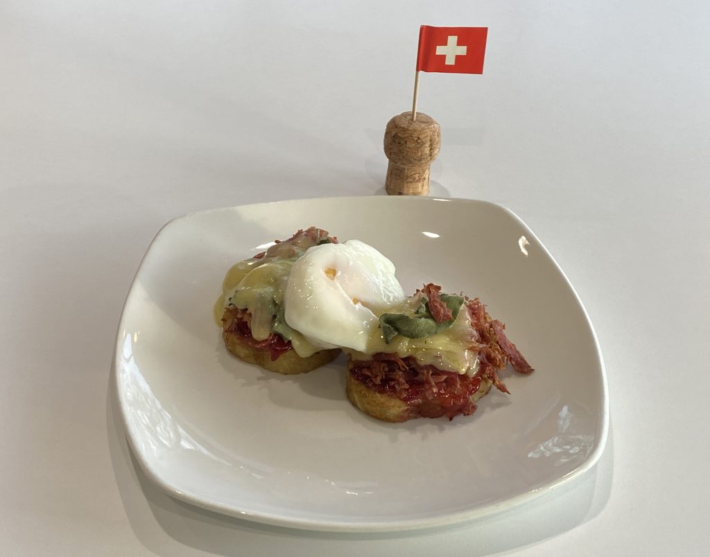 Two potato röstis with served with ham hock, melted cheese, sage leaves and a poached egg. The plate sits on a table next to a Swiss cocktail stick flag.