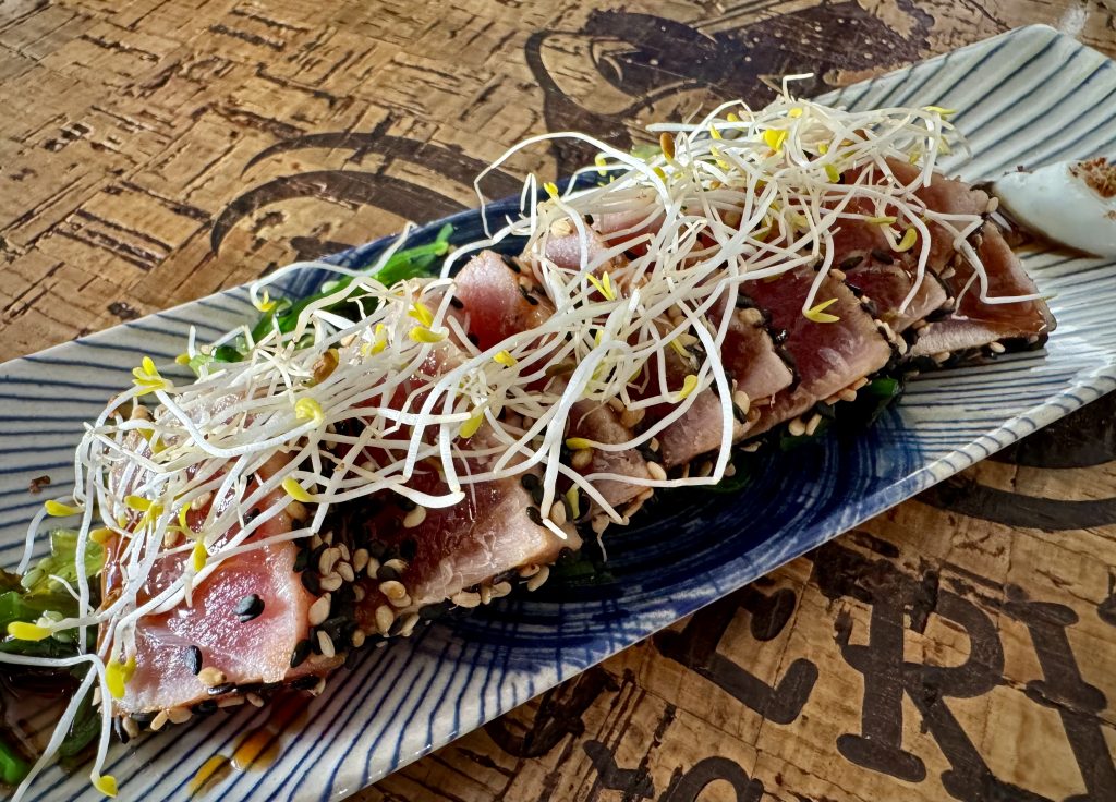 A close-up of nine tuna tataki slices stacked on angle on a blue plate. They are garnished with cress.