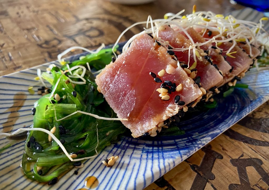 Five slices of tuna tataki, stacked on an angle on a long blue plate. Wakame seaweed, toasted sesame seeds and cress accompany it.