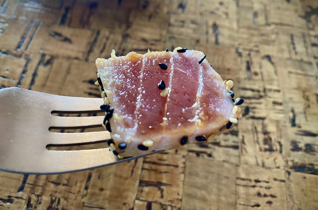 A close-up of a slice of tuna tataki on the end of a fork. Its layers are visible, as is the toasted sesame coating.