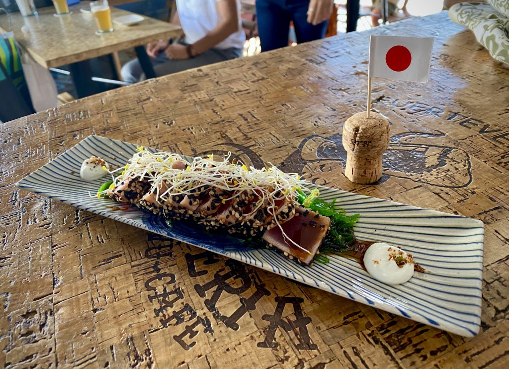 A plate of tuna tataki, coated in toasted sesame seeds and garnished with daikon radish sprouts. It sits on a table next to a Japanese cocktail stick flag.