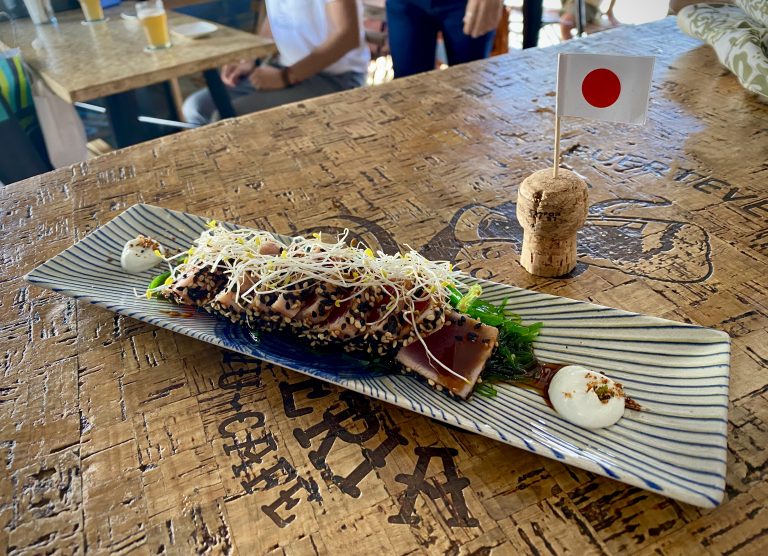 A plate of tuna tataki, coated in toasted sesame seeds and garnished with daikon radish sprouts. It sits on a table next to a Japanese cocktail stick flag.