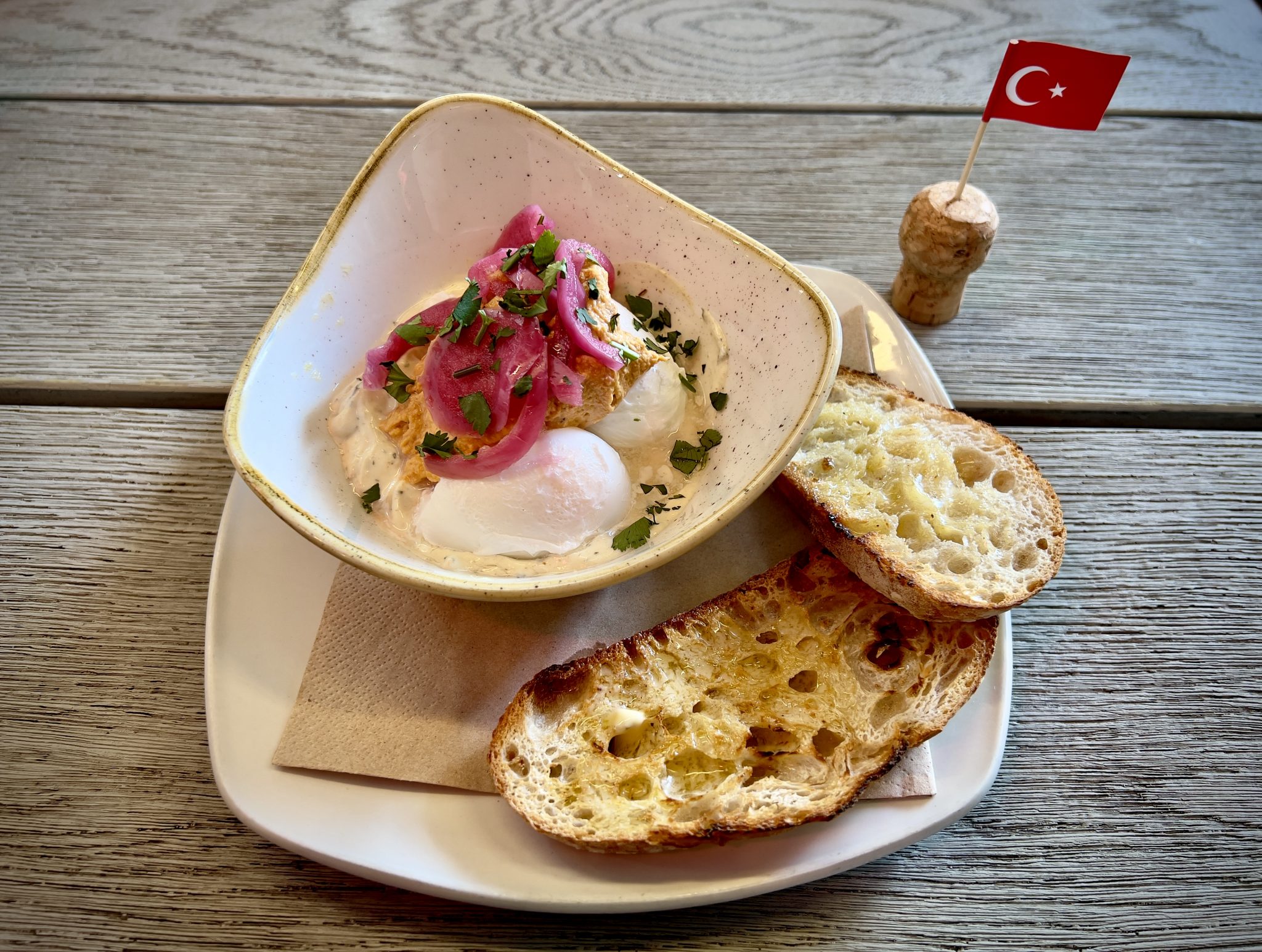 Çılbır (Turkish Eggs) served in a bowl, next to two slices of buttered sourdough toast on a white plate. The plate is on a wooden table, next to a Turkish cocktail stick flag and cork.